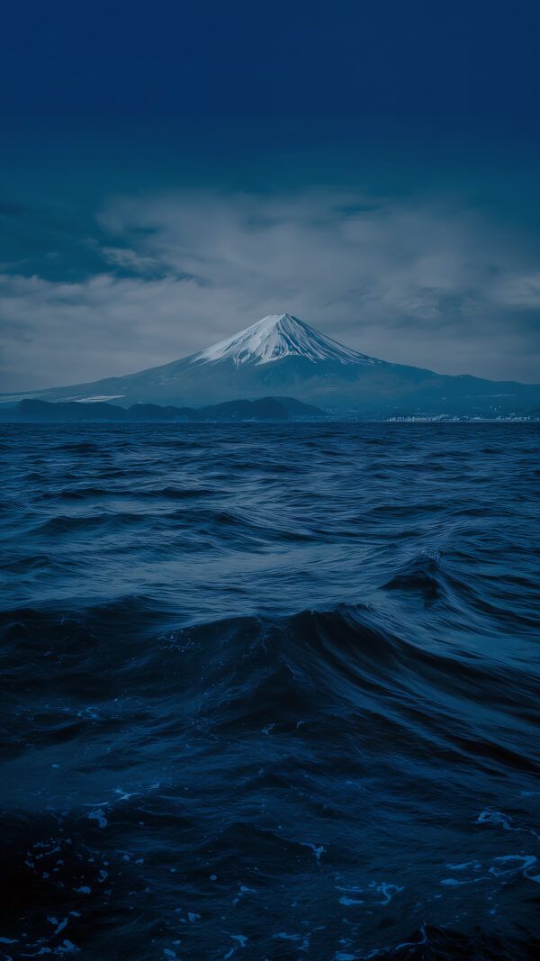 The snow-capped Mount Fuji seen across a dark, choppy body of water under a cloudy, dramatic sky.