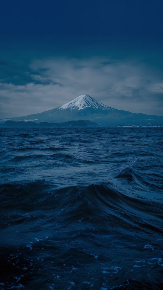 The snow-capped Mount Fuji seen across a dark, choppy body of water under a cloudy, dramatic sky.
